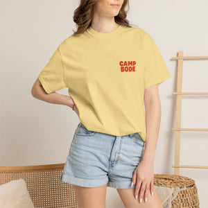 A woman stands in a beige room with minimal decor, wearing the Printify BODE Summer Camp T-Shirt 2026 "Throwback Sunset" (Unisex Comfort Colors 1717) and denim shorts.