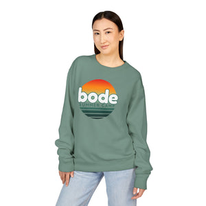 Person wearing the Printify 2026 Bode Summer Bootcamp Crewneck Sweatshirt GDH400, featuring a sunset graphic, stands facing the camera with one hand in their pocket.