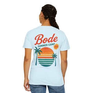 Person facing away, wearing the Printify BODE Summer Camp T-Shirt 2026 — "Throwback Sunset" (Unisex Comfort Colors 1717) in light blue with "Bode Summer Camp 2026" and a sunset, palm trees, and stripes graphic on the back.