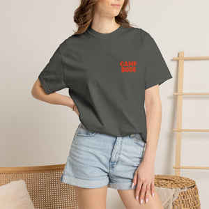 A woman stands indoors by a wooden chair and ladder shelf, wearing the Printify BODE Summer Camp T-Shirt 2026 "Throwback Sunset" (Unisex Comfort Colors 1717) in dark gray with light denim shorts.