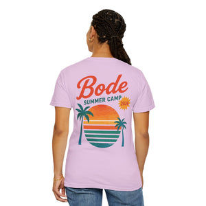 A person wears the Printify BODE Summer Camp T-Shirt 2026 "Throwback Sunset," a light purple, garment-dyed unisex tee featuring palm trees and a sunset graphic on the back.