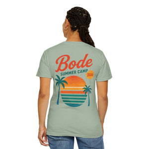 A person faces away, wearing the Printify BODE Summer Camp T-Shirt 2026 "Throwback Sunset" (Unisex Comfort Colors 1717), featuring a light green heavyweight cotton fabric with a sunset and palm trees graphic on the back.