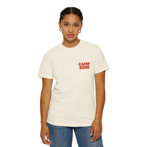 A person stands facing forward wearing the Printify BODE Summer Camp T-Shirt 2026 "Throwback Sunset," a cream Comfort Colors 1717 shirt with red "CAMP BODE" text on the left chest, paired with blue jeans.