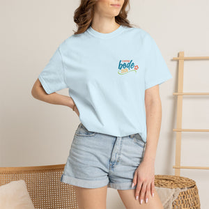 Person wearing a light blue Printify BODE Summer Camp T-Shirt 2026—"Life's Better Together"—Unisex Comfort Colors 1717, paired with denim shorts, stands indoors with one hand on their hip.