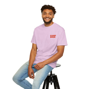 A man in a light purple BODE Summer Camp T-Shirt 2026 "Throwback Sunset" by Printify sits on a black stool, smiling in light blue jeans against a white background.