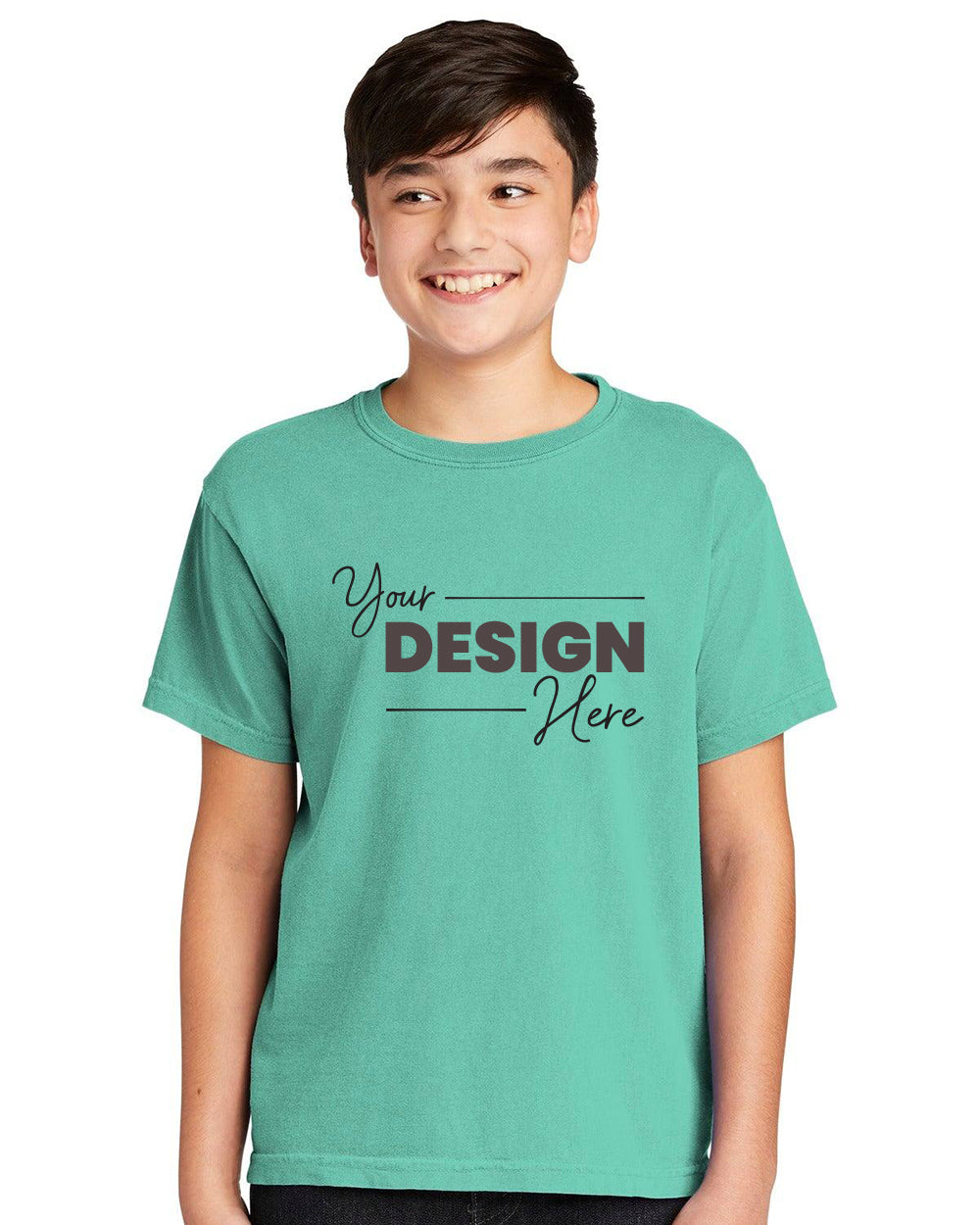 A boy wears a teal Comfort Colors Youth Ring Spun T-Shirt 9018 featuring black "Your Design Here" text, offering a vintage look. Crafted from ring-spun cotton, it combines comfort and style.