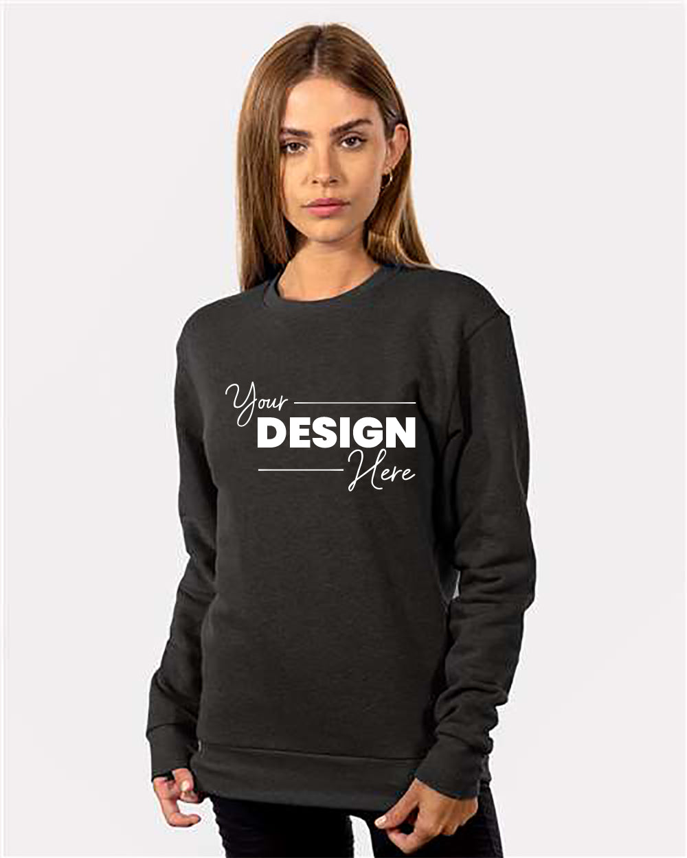 A woman is wearing the Next Level Unisex Malibu Crewneck Sweatshirt in dark grey, with the words "Your Design Here" printed on the front. Made by Next Level from heavyweight fabric, this piece features a relaxed fit and a tear-away label for added comfort. She stands against a plain white background.