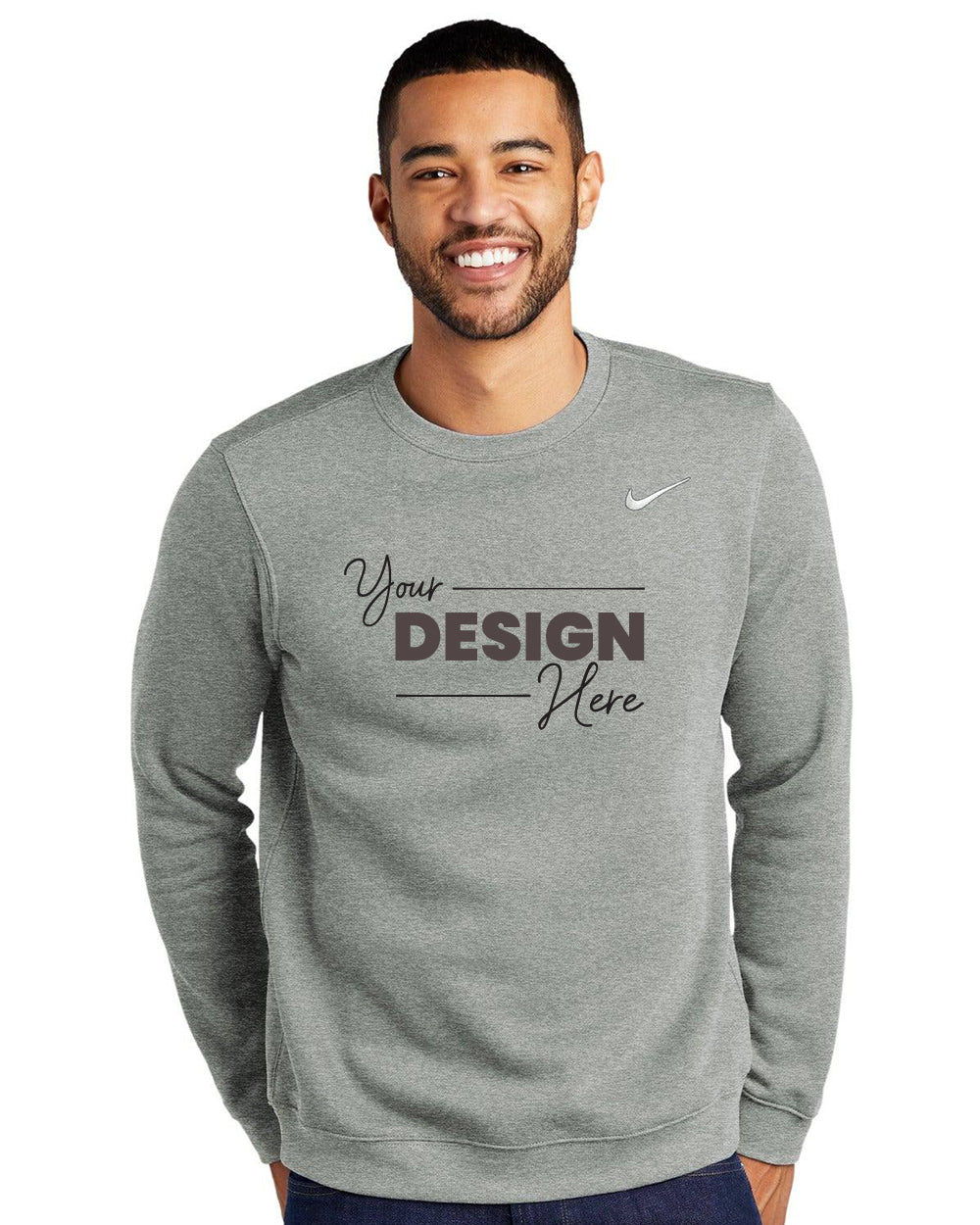 A smiling person wears a gray Nike Club Fleece Crewneck Sweatshirt CJ1614 with "Your Design Here" on the front and an embroidered Swoosh, set against a white background.