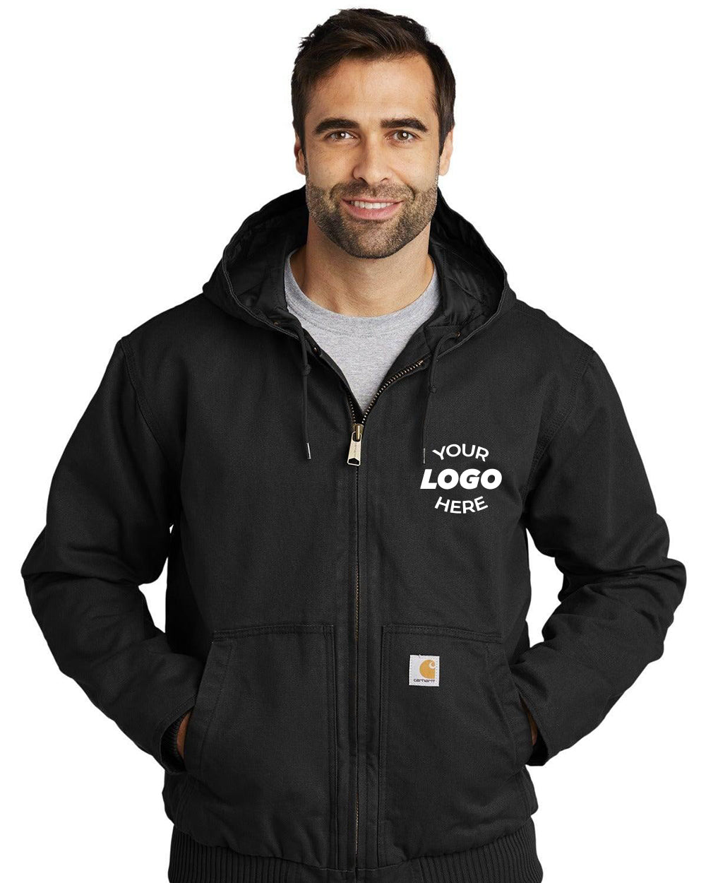A man wearing a black Carhartt Washed Duck Active Jacket CT104050, known for its durability, with "Your Logo Here" on the left chest, stands with hands in pockets like a rugged Carhartt adventurer.