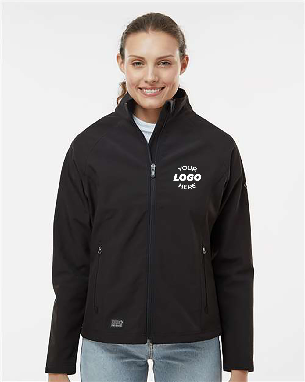A woman stands smiling in a black DRI DUCK Women's Contour Soft Shell Jacket featuring the text "Your Logo Here." She is wearing light blue jeans and a white turtleneck underneath the soft shell jacket, which is both wind and water-resistant.