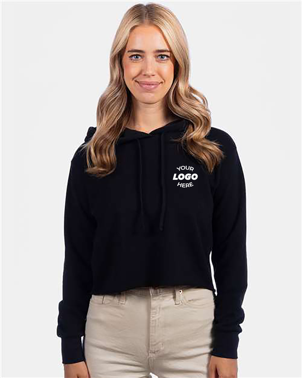A woman with long blonde hair wears a black Next Level Women's Unisex Laguna Sueded Hoodie Sweatshirt that has "Your Logo Here" written on it and beige pants. She is standing against a plain white background.