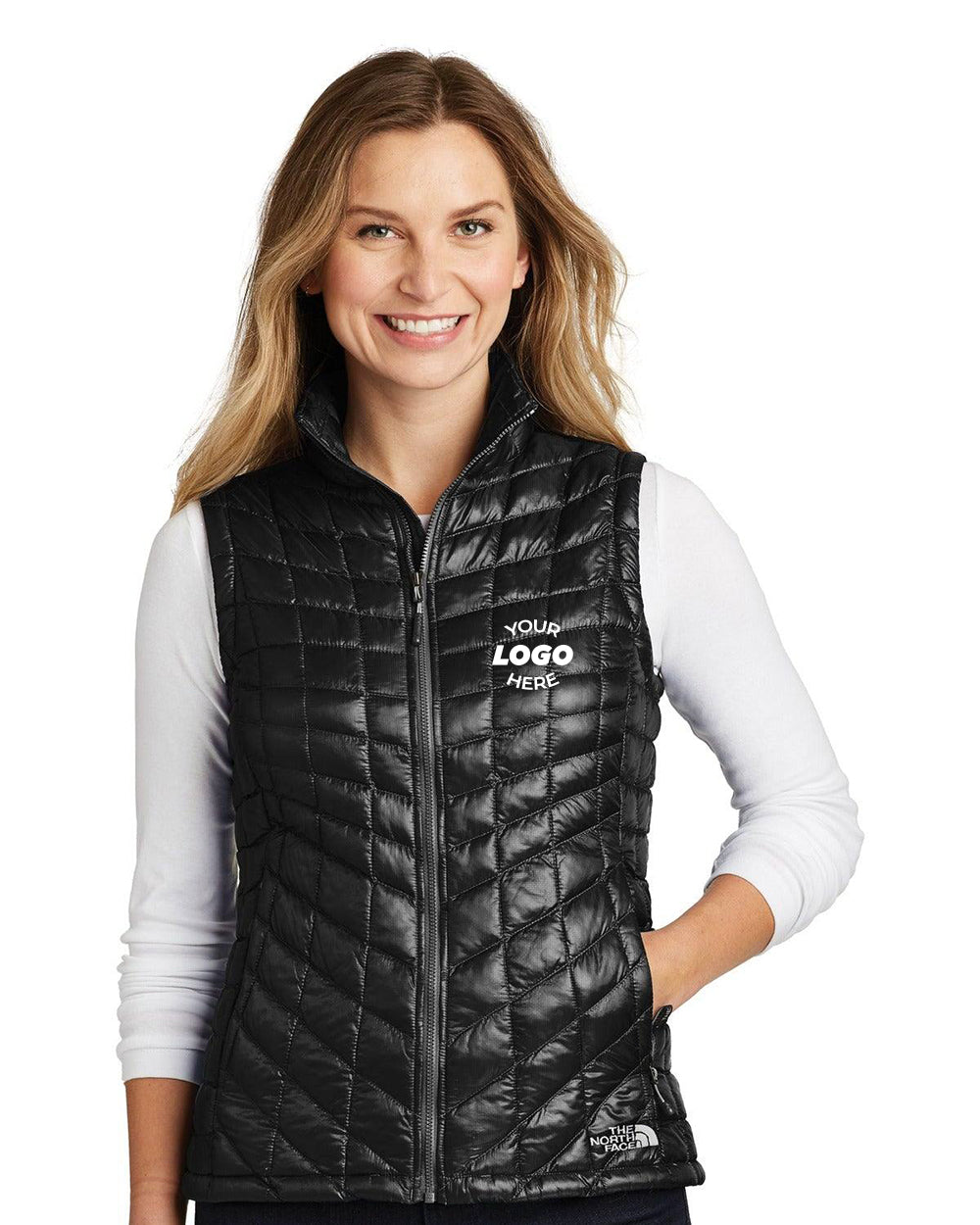 A woman smiles at the camera, wearing The North Face Ladies ThermoBall Trekker Vest NF0A3LHL in black with "Your Logo Here" on the chest, layered over a white long-sleeve shirt.