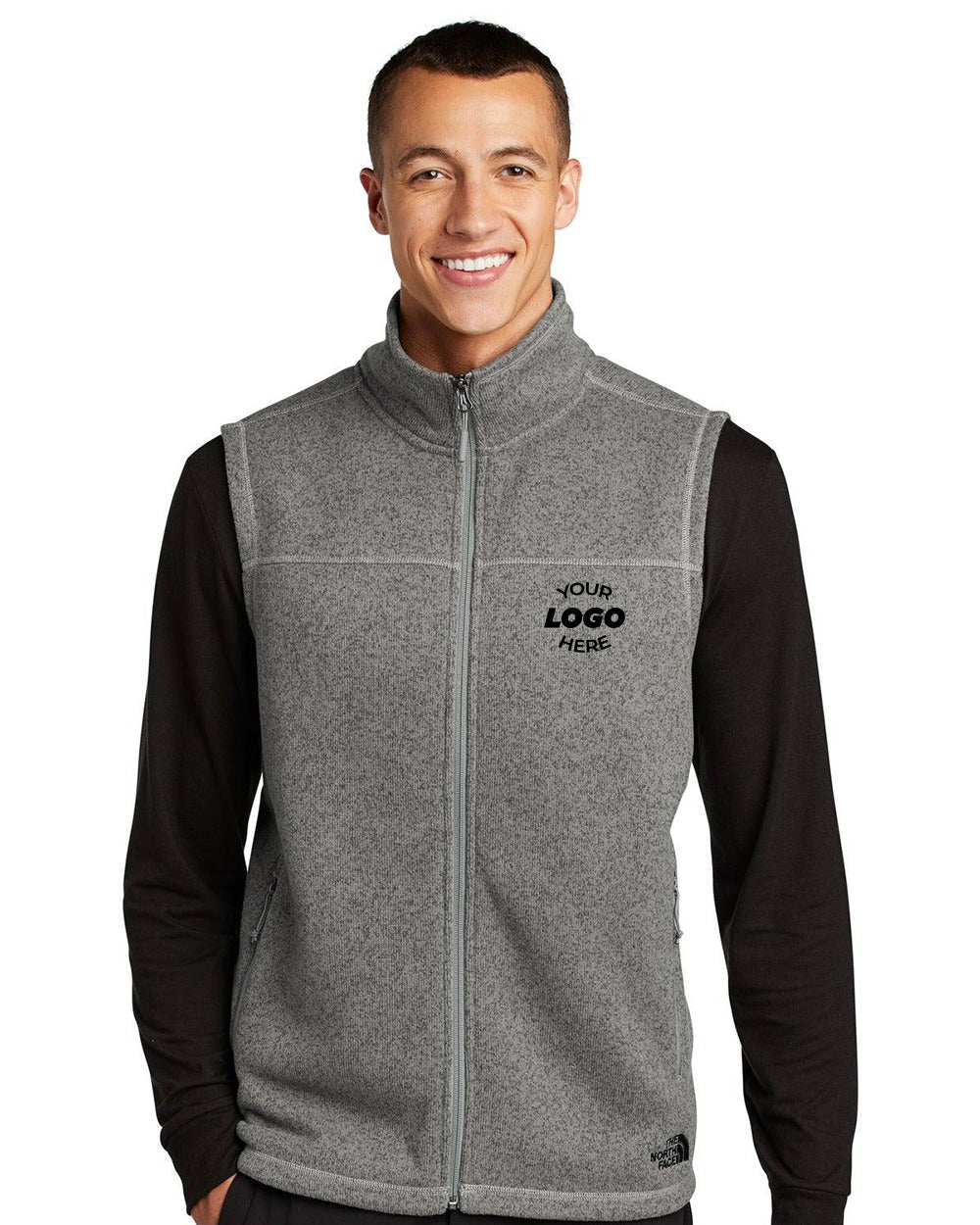 A person smiles in a gray The North Face Sweater Fleece Vest NF0A47FA and a black long-sleeve shirt. "Your Logo Here" stands out on the vest's cozy polyester sweater knit, echoing The North Face style.