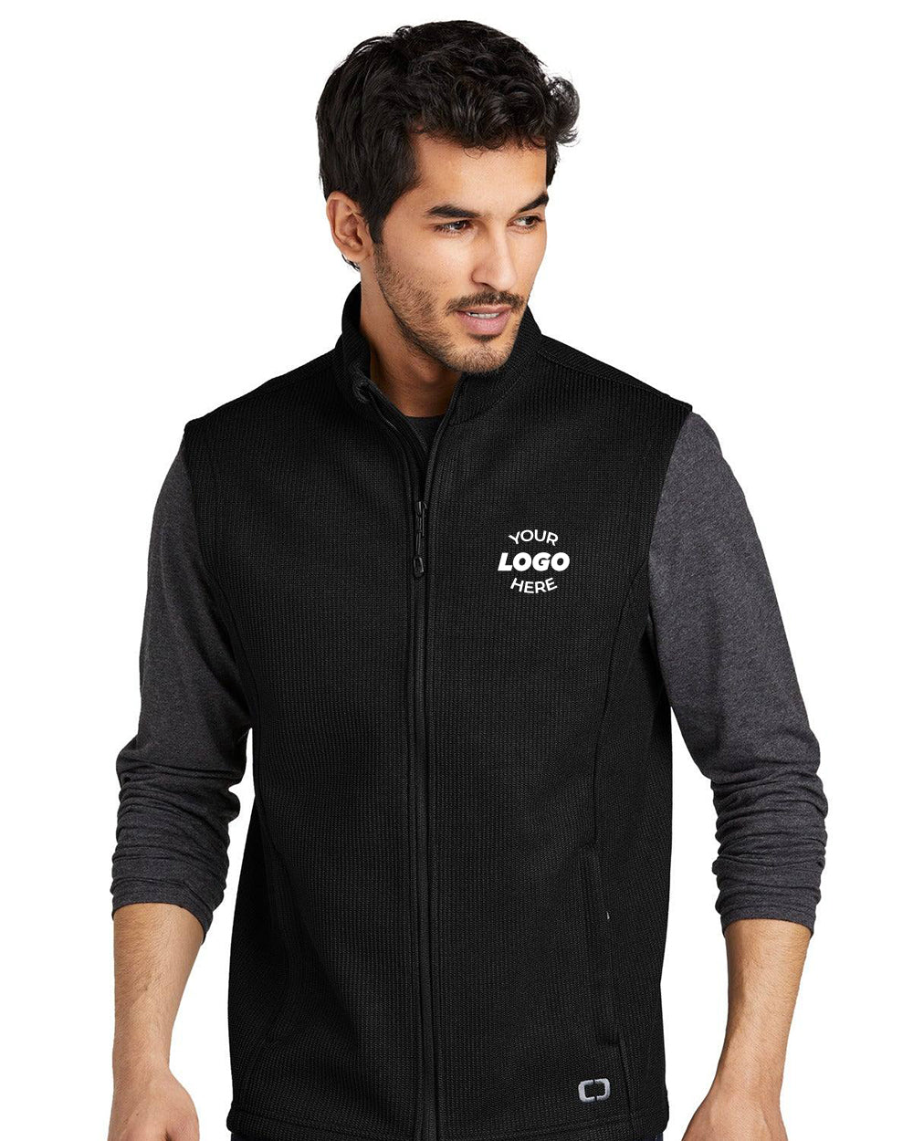 Man wearing an OGIO Grit Fleece Vest (OG730) in black, featuring an OGIO zipper pull and "YOUR LOGO HERE" text on the left chest, layered over a long-sleeve gray shirt.