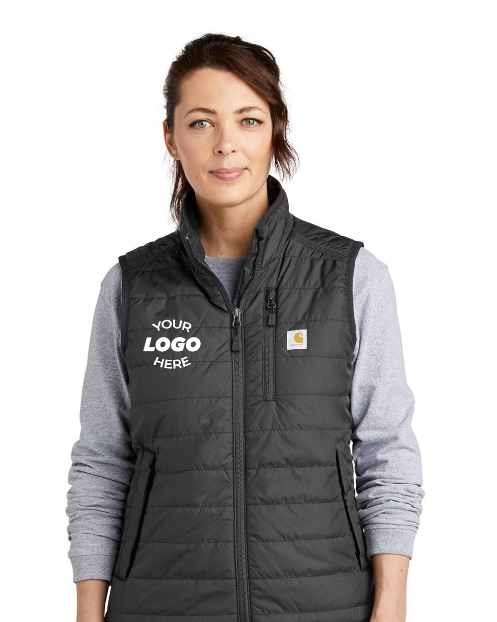 A woman wearing the Carhartt Women's Gilliam Vest CT104315—a gray, wind-resistant quilted vest with a small Carhartt logo and "Your Logo Here" area—over a light gray long-sleeve shirt.