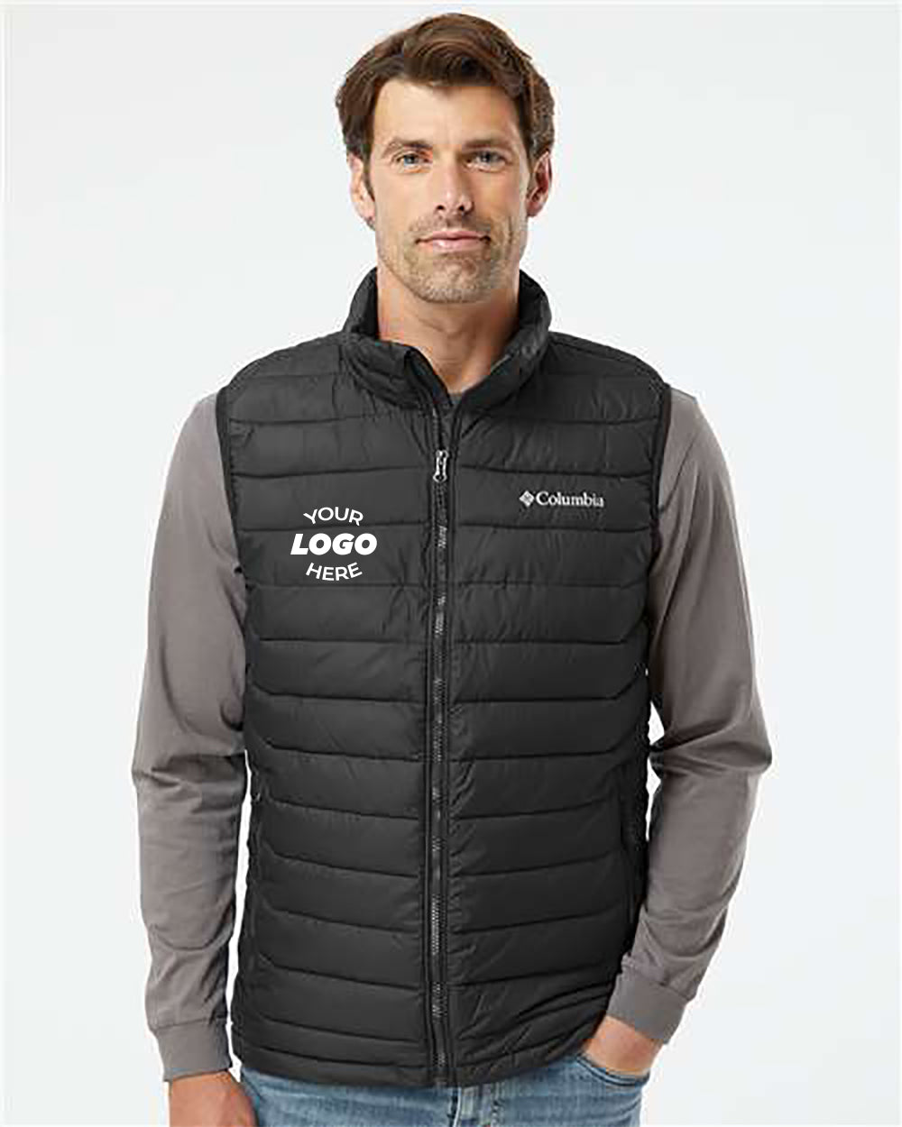 A man poses wearing a black Columbia Powder Lite Vest over a grey long-sleeve shirt. The vest, ideal for durable workwear, displays a "Your Logo Here" placeholder on the left side and includes Thermarator™ insulation for added warmth.