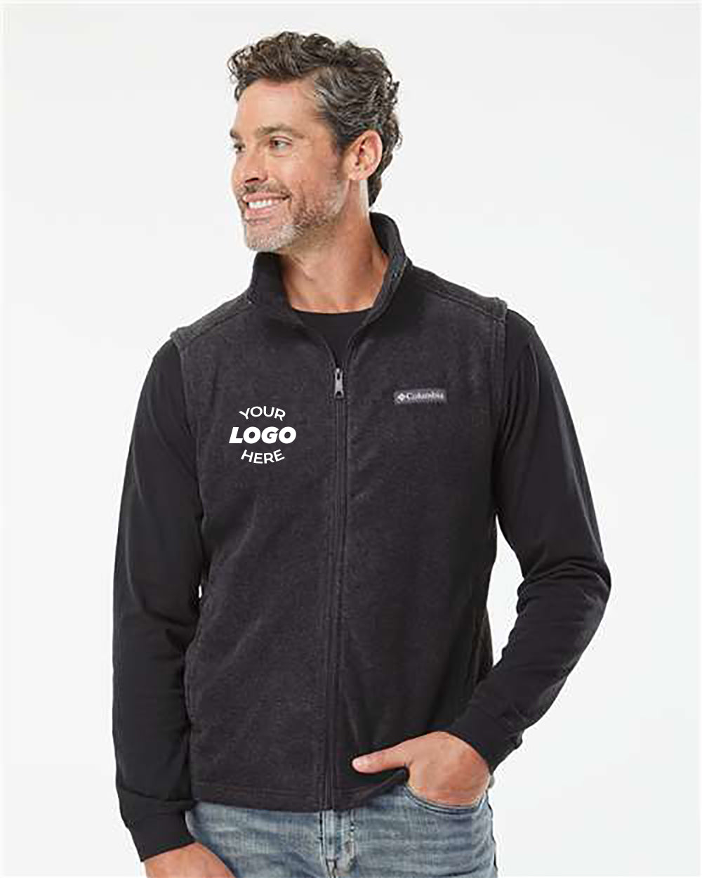 A man with short, curly hair wears a black Columbia Steens Mountain Fleece Vest from Columbia over a black long-sleeve shirt. The polyester fleece vest features "YOUR LOGO HERE" text on the left chest area. He is looking to the side, smiling, and ready for outdoor activities.