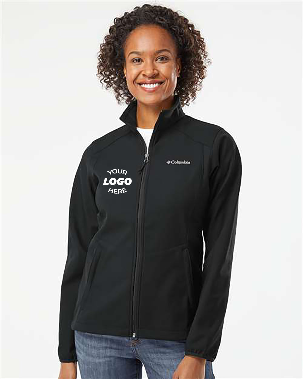 A person wearing a Columbia Womens Kruser Ridge Softshell Jacket in black, with "Your Logo Here" printed on the front, is standing and smiling against a plain background. The jacket, from Columbia, features a comfort stretch design and water-and-wind-resistant properties, making it perfect for any weather conditions.