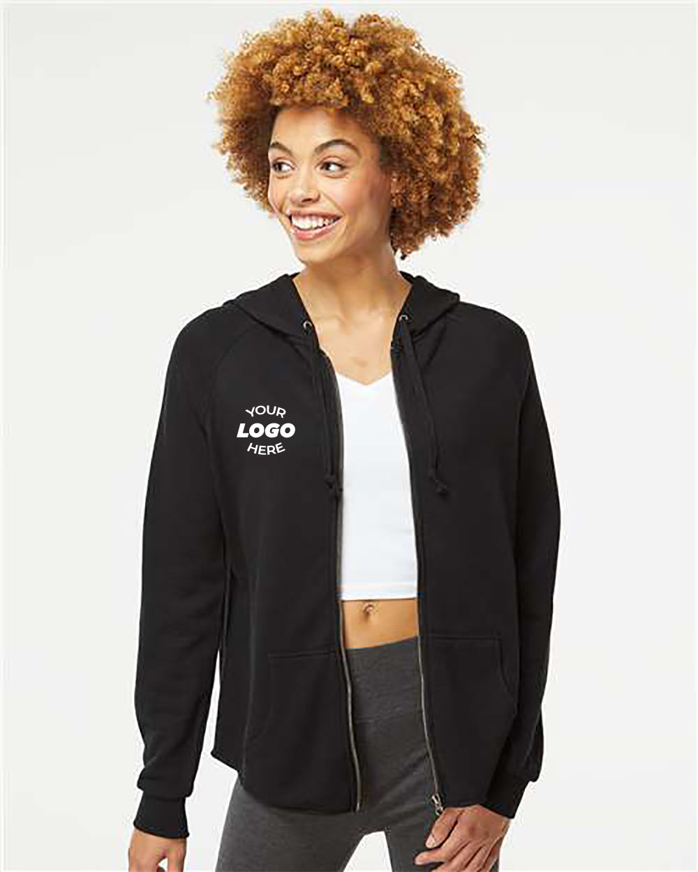 A person smiles while wearing an Independent Trading Co. Women's California Wave Wash Full-Zip Hoodie Sweatshirt with a white logo on the left chest, a white top, and gray pants, standing against a plain background.