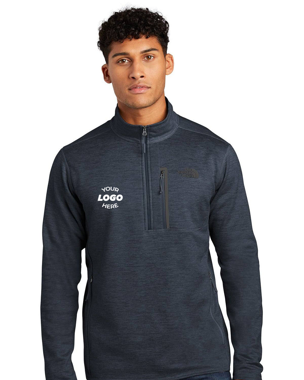 A man wears The North Face Skyline 1/2-Zip Fleece Pullover (NF0A7V63) in dark gray, made from recycled polyester, featuring "YOUR LOGO HERE" on the left chest and a vertical zippered pocket on the right chest.