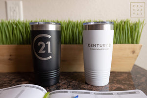 Two Kodiak Coolers tumblers engraved, one black and one white, with "Century 21" logos, displayed on a desk beside a planner and artificial grass.