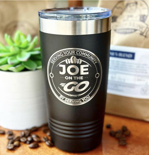 A Kodiak Coolers black insulated tumbler with the "Joe On The Go" logo sits on a wooden surface, surrounded by coffee beans, a succulent, and a coffee bag. Ideal as an engraved tumbler or custom corporate promotional gift for those valuing quality and style. Buy now: no setup fees & limited time offer!.