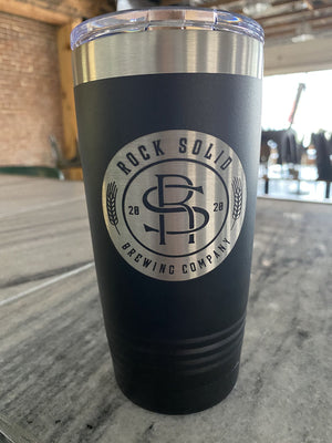 A black insulated tumblers from Kodiak Coolers, custom-engraved with the logo of Rock Solid Brewing Company featuring stylized letters "RS" and the year 2020, on a wooden table in a room.