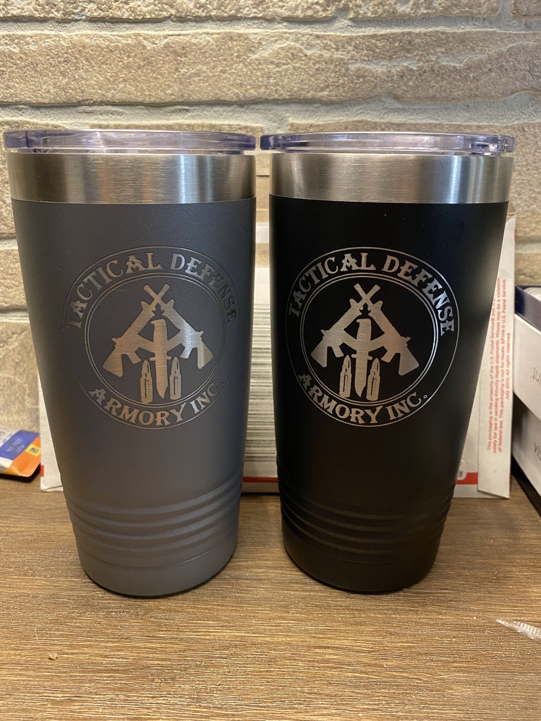 Two large, Custom Tumblers 20 oz with your Logo or Design Engraved - Special Bulk Wholesale Pricing - Pack of 48 Pieces - 1 Color - $14.56 Each from Kodiak Coolers in gray and black, each with a laser-engraved logo featuring a shield, two rifles, and two bullets, resting on a table against a brick wall background.