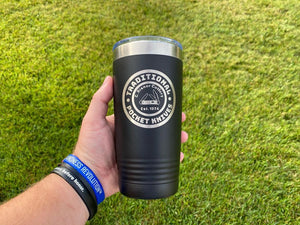A person's hand holding a black insulated tumbler with "traditional iron works" and "pocket ninja" printed on it, against a grassy background, potentially ideal for corporate promotional gifts by Kodiak Coolers.