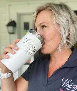 A woman with short blonde hair drinking from a white Kodiak Coolers custom tumbler engraved with a cursive logo, wearing a navy polo shirt with a pink logo.