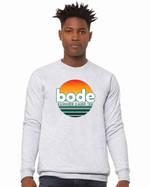 A person wears a light gray Bode Summer Bootcamp "OG Logo" unisex crewneck BC Fleece Raglan Sweatshirt 3901 by designerapp, featuring a green and orange logo on the front and made from pre-shrunk Airlume cotton for comfort.