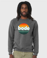 Man wearing a gray designerapp Bode Summer Bootcamp "OG Logo" unisex crewneck BC Fleece Raglan Sweatshirt 3901 featuring a colorful logo and sunset graphic, against a light plain background.