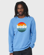 A person wearing a light blue designerapp Bode Summer Bootcamp "OG Logo" unisex crewneck BC Fleece Raglan Sweatshirt 3901, featuring a sunset graphic on the front and crafted with soft fleece using Direct to Garment printing.
