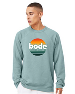 Someone wearing a light blue designerapp Bode Summer Bootcamp "OG Logo" unisex crewneck BC fleece raglan sweatshirt 3901 featuring a retro-style logo and sunset graphic on the front.