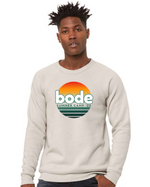 A person wearing a light-colored designerapp Bode Summer Bootcamp "OG Logo" unisex crewneck fleece raglan sweatshirt (3901) with a sunset-inspired front graphic.