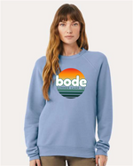 A woman wears the designerapp Bode Summer Bootcamp "OG Logo" unisex crewneck BC fleece raglan sweatshirt 3901 in light blue, featuring a sunset logo on soft, pre-shrunk Airlume combed and ring-spun cotton.