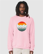 A man wears a light pink designerapp Bode Summer Bootcamp "OG Logo" unisex crewneck BC Fleece Raglan Sweatshirt 3901 featuring a sunset graphic, crafted from soft Airlume combed and ring-spun cotton with DTG printing.