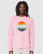 A person wearing the designerapp Bode Summer Bootcamp "OG Logo" Unisex Crewneck BC Fleece Raglan Sweatshirt 3901 in light pink, made from soft Airlume combed and ring-spun cotton with a sunset logo on the front.
