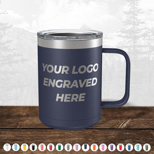 A navy blue insulated tumbler with a handle, custom printed with the text "your logo engraved here," on a wooden surface against a blurred forest background. TODAY ONLY - Hump Day Sale - Your Logo Engraved on Drinkware - Single Side Engraving Included in Price - Slider Lids Included by Kodiak Coolers.
