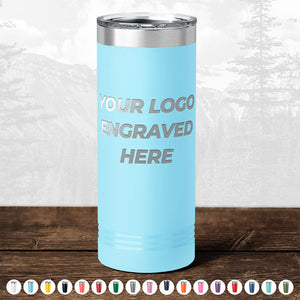 A light blue insulated tumbler with "your logo engraved here" text, displayed on a wooden table against a blurred forest background, perfect as a promotional gift from Kodiak Coolers.