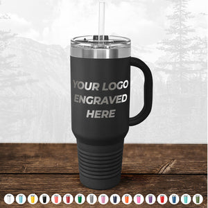 Black insulated travel mug with a handle and a straw, featuring the text "TODAY ONLY - Hump Day Sale - Your Logo Engraved on Drinkware - Single Side Engraving Included in Price - Slider Lids Included" on a wood surface against a blurred forest background from Kodiak Coolers.