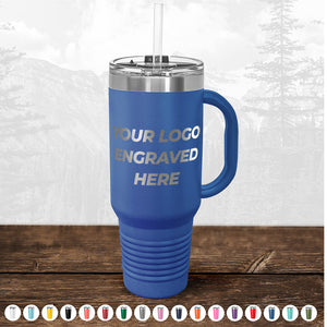 A blue insulated tumbler with a handle and straw, custom printed with the text "your logo engraved here" on its side, set on a wooden surface against a faded forest background from Kodiak Coolers.