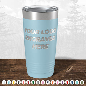 A light blue insulated tumbler from Kodiak Coolers with "your logo engraved here" text, displayed on a wooden table against a blurred forest background. Various color options for these promotional gift mugs are shown below.