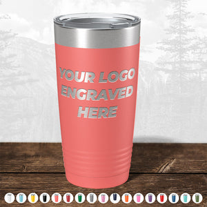 A coral-colored Kodiak Coolers insulated tumbler with a silver lid and "your custom logo here" text, displayed on a wooden surface against a misty forest background.