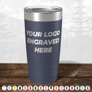 A navy blue insulated tumbler from Kodiak Coolers with "your custom logo here" text, displayed on a wooden surface with a blurred forest background.