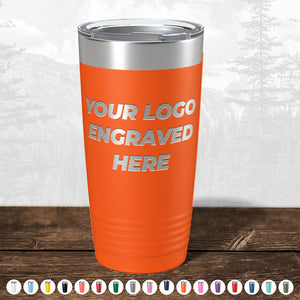 Orange insulated tumbler from Kodiak Coolers with "your custom logo engraved here" text, displayed on a wooden surface against a blurred forest background.
