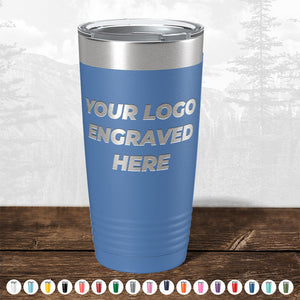 A blue insulated tumbler with "your custom logo here" on it, displayed on a wooden surface against a blurred forest background from Kodiak Coolers.