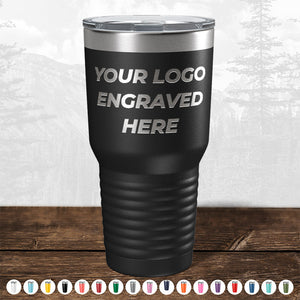 A customizable Kodiak Coolers black travel mug with "your logo engraved here" text, perfect as a promotional gift, displayed on a wooden surface with a blurry forest background.