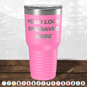 A pink insulated tumbler labeled "Your Logo Engraved Here" is displayed on a wooden surface with a mountain backdrop. Kodiak Coolers' custom tumblers are ideal corporate gifts, available in various colors with bulk wholesale pricing and no setup fees.