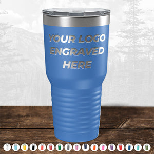 Blue insulated tumbler with Kodiak Coolers "your custom logo here" text, displayed on a wooden surface against a blurred forest background.